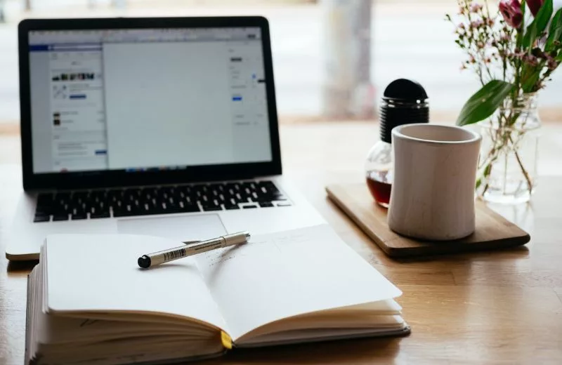 Laptop sitting open on desk next to a cup of coffee, flowers, and a journal