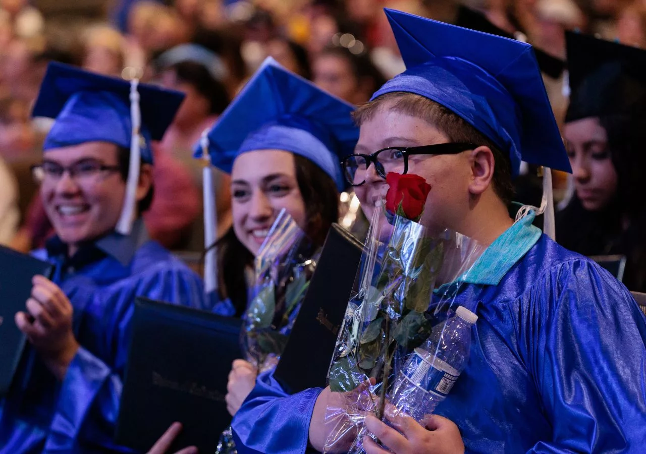 Student sitting with other graduates, holding a rose and their diploma