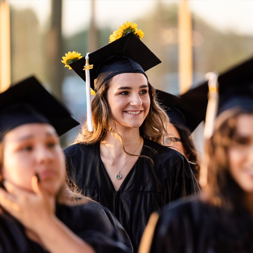 A graduate wearing a cap and gown, smiling as she enters her graduation ceremony with fellow graduates.