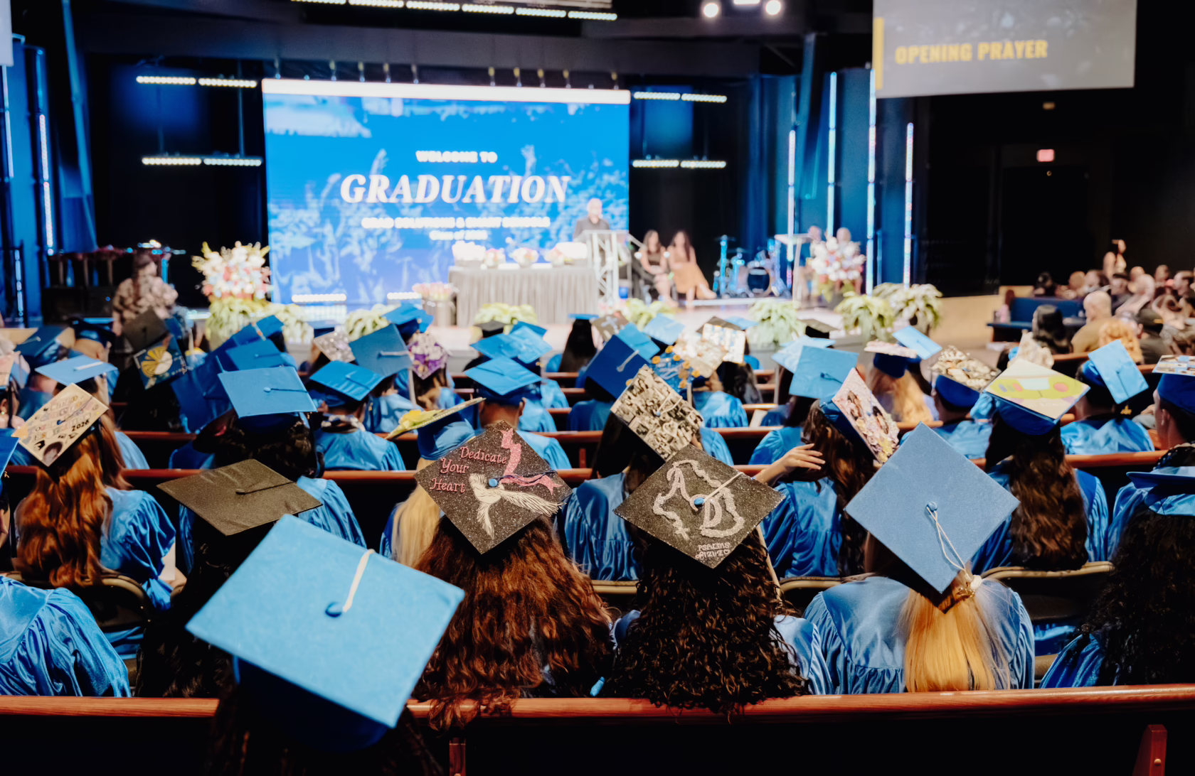 Rows of graduates in blue caps and gowns sit facing a stage during a high school graduation ceremony, with many decorated grad caps visible in the audience.