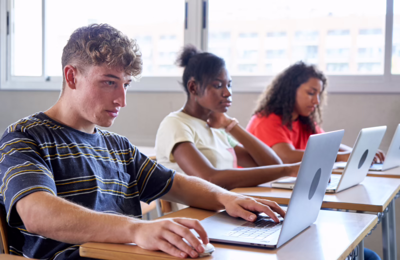 Side view of teenage students in a classroom studying with technological equipment, using laptops.