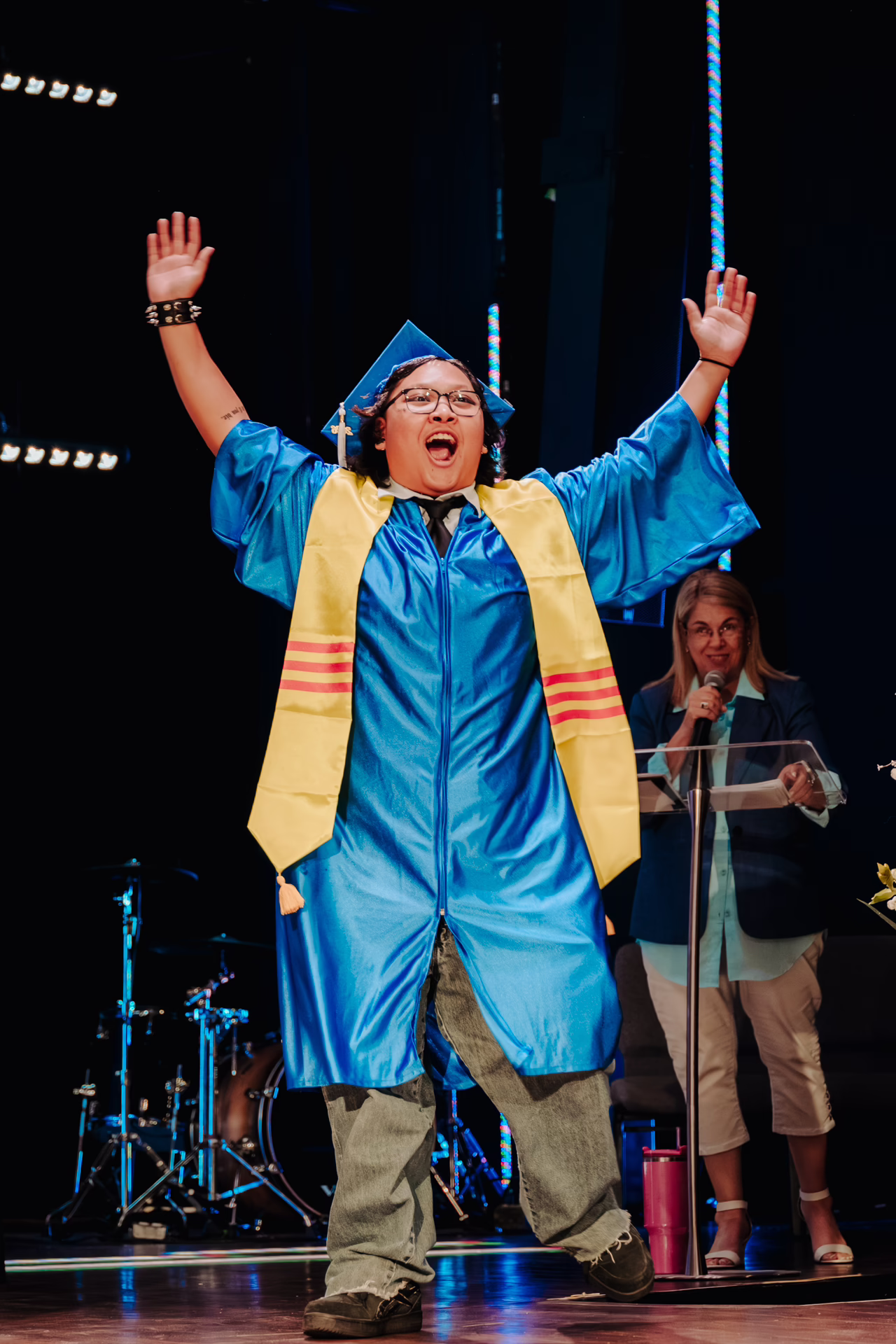 Graduation ceremony participant in a blue cap walks across a stage with both arms raised.