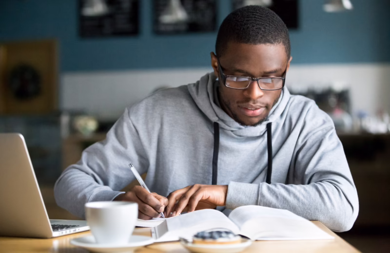 Focused millennial african american student in glasses making notes writing down information from book in cafe preparing for test or exam, young serious black man studying or working in coffee house