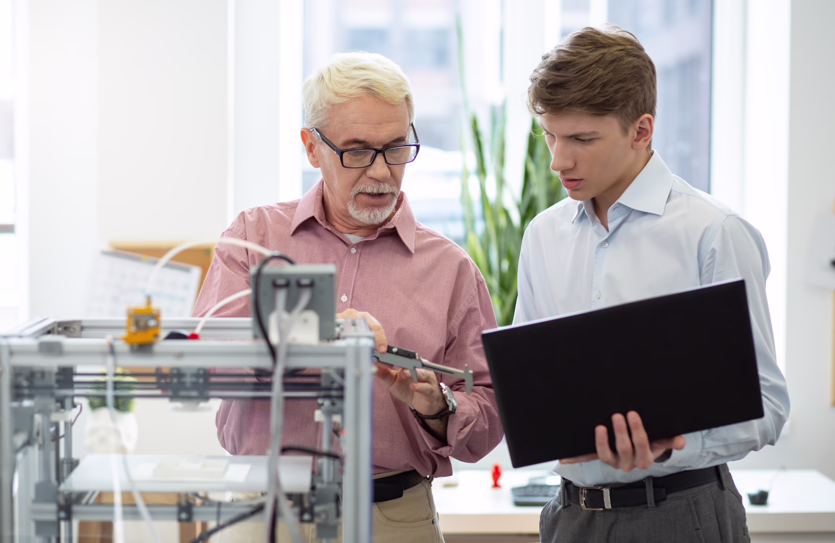 Two people stand beside a desktop 3D printer in a bright workshop. One person holds a laptop while the other uses a measuring tool to inspect a component. Large windows and a plant are visible in the background.