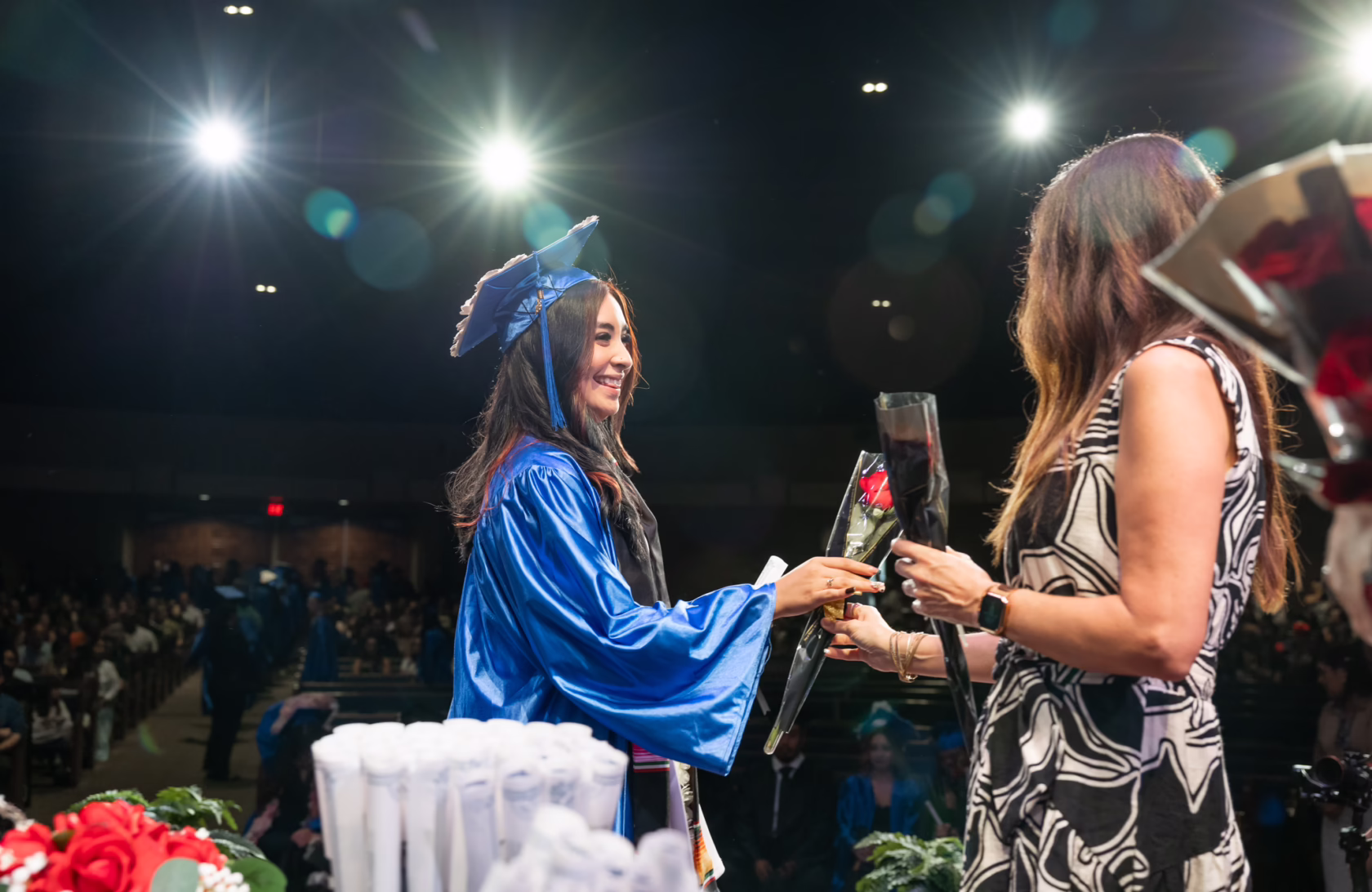 Graduate wearing blue cap and gown walks across the stage during a ceremony to receive a rose