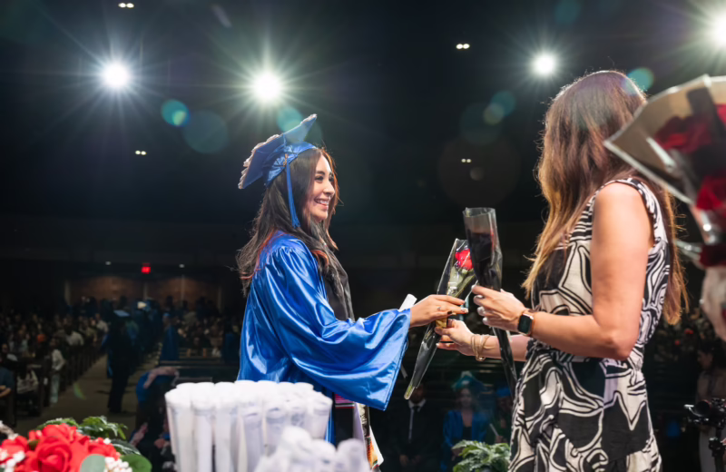 Graduate wearing blue cap and gown walks across the stage during a ceremony to receive a rose